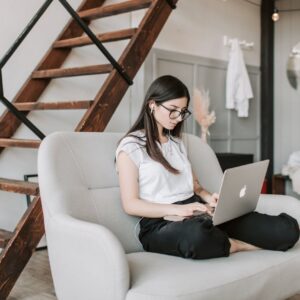 Young female in eyeglasses and casual clothes sitting barefoot on sofa in modern apartment with wooden staircase while wearing earbuds and watching movie on netbook
