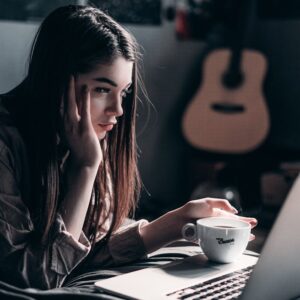 Woman lying on bed, focused on laptop with a coffee cup in hand, embracing remote work lifestyle.