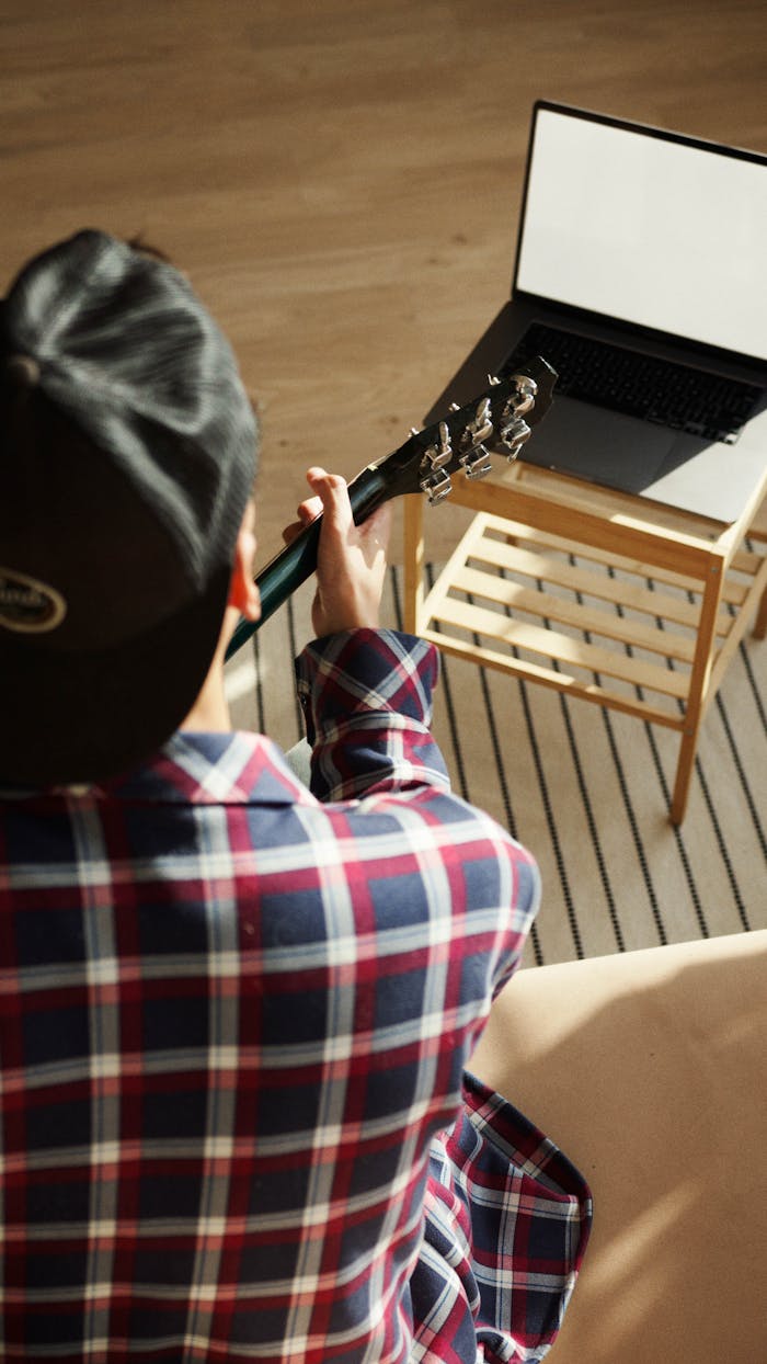 gallery-01 A man in plaid shirt strums a guitar next to a laptop, enjoying music indoors.