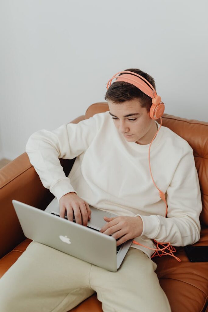 Teen using laptop with headphones, relaxed on a couch at home.