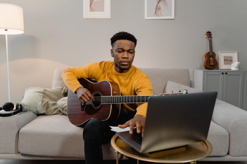 A man in a yellow sweater plays guitar while using a laptop in a cozy living room.