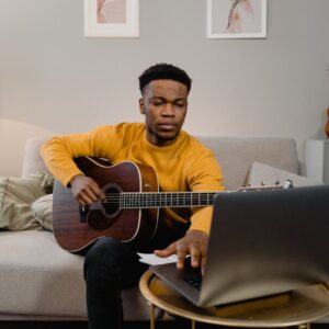 A man in a yellow sweater plays guitar while using a laptop in a cozy living room.