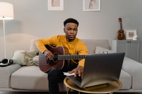A man in a yellow sweater plays guitar while using a laptop in a cozy living room.