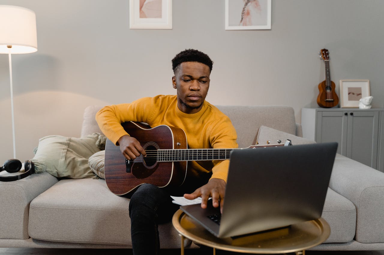 services-02 A man in a yellow sweater plays guitar while using a laptop in a cozy living room.