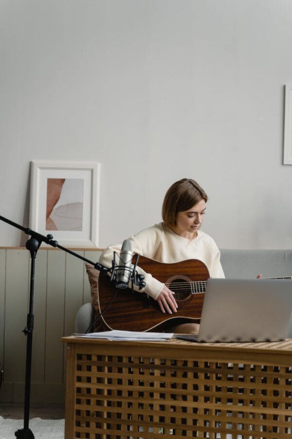 Adult woman playing guitar with laptop for online music learning. Indoors, cozy setting.