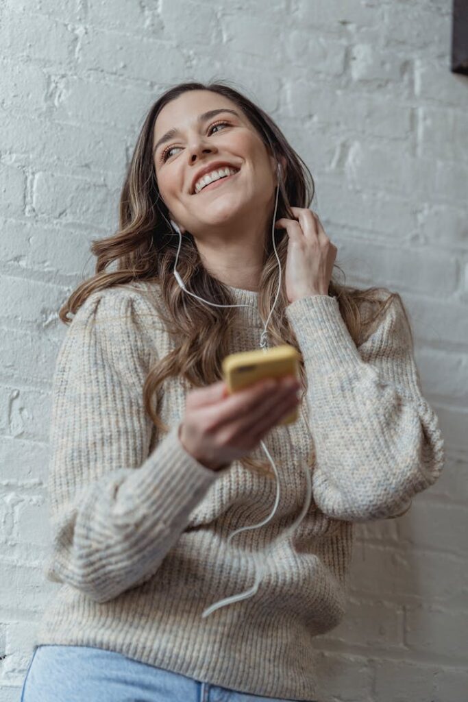 Cheerful young female in sweater browsing mobile phone and listening to favorite music in earphones against brick wall