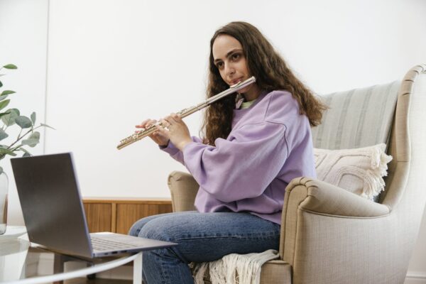 Woman wearing a purple sweater plays flute while attending an online music lesson from home.